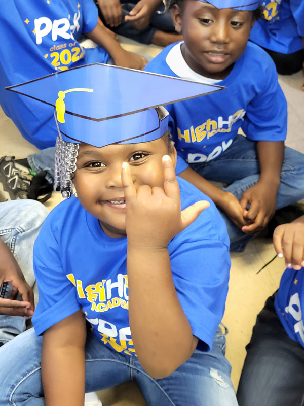 Child in graduation cap smiling and gesturing a 'love' sign.