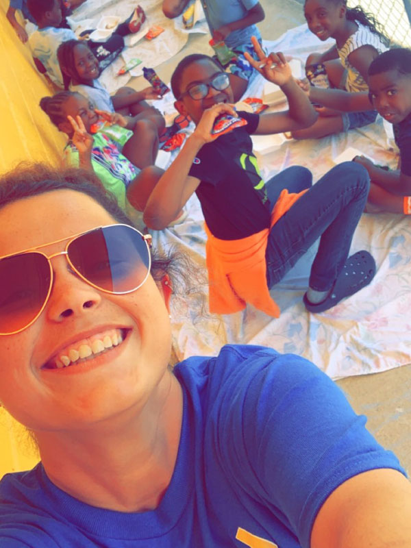 Group of children and an adult smiling and making hand signs on a picnic blanket.