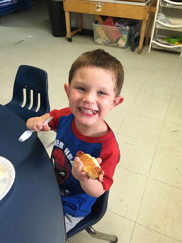 Child eating a hotdog at a table with a big smile.