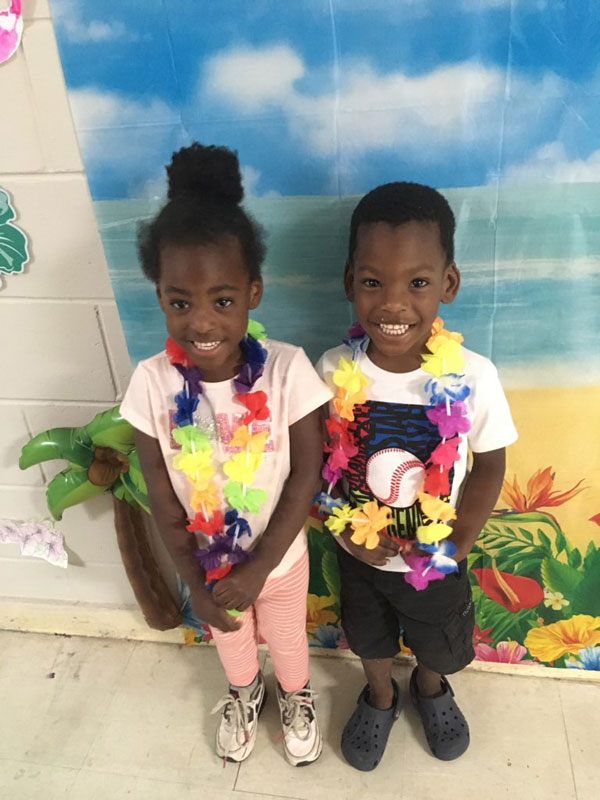 Two children wearing leis and smiling in front of a tropical backdrop.